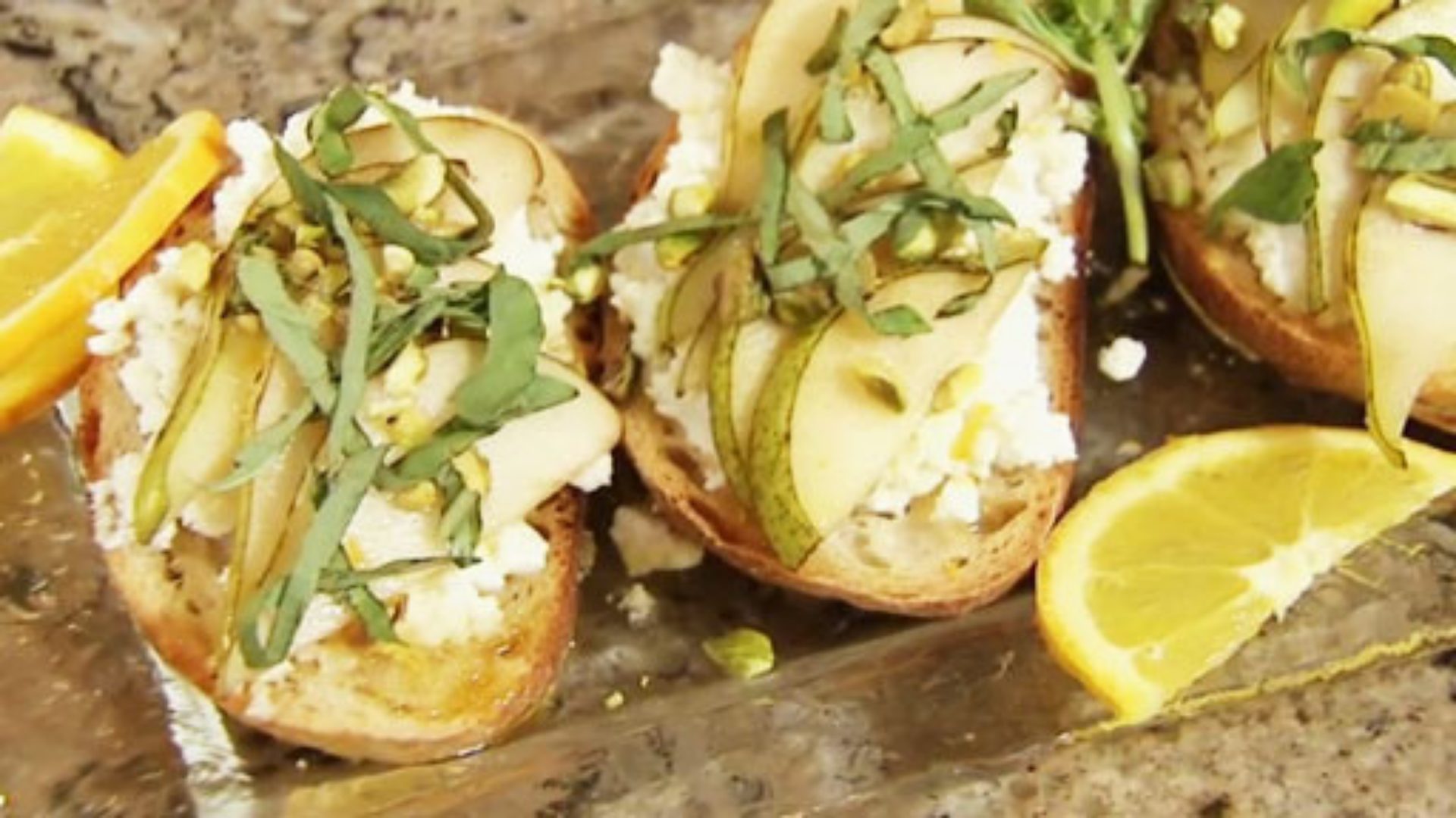 Three slices of toasted bread topped with slices of pear, rich ricotta cheese, and herbs, with a side of sliced lemon. 