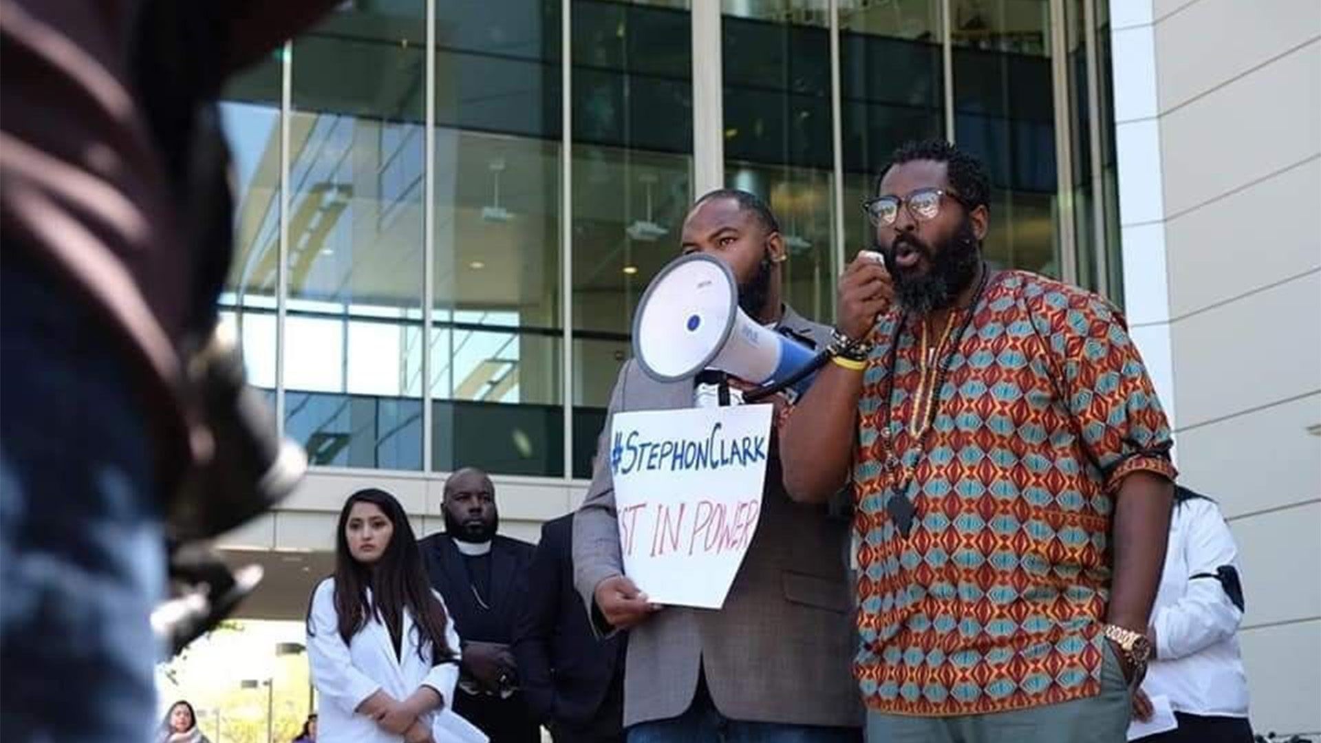 Two men stand in the middle of a crowd, one is holding a speaker and the other a microphone.